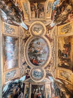 A beautifully ornate ceiling featuring intricate frescoes and detailed paintings framed by elaborate gold borders. The central image depicts mythological or religious figures, surrounded by additional vignettes showcasing various scenes involving people and classical architectural elements. The artwork displays a harmonious blend of rich colors and Renaissance influences.