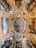 A beautifully ornate ceiling featuring intricate frescoes and detailed paintings framed by elaborate gold borders. The central image depicts mythological or religious figures, surrounded by additional vignettes showcasing various scenes involving people and classical architectural elements. The artwork displays a harmonious blend of rich colors and Renaissance influences.