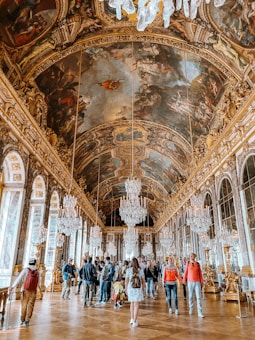 An opulent hall with a high ornate ceiling covered in elaborate paintings. Crystal chandeliers hang from the ceiling, reflecting light in the grand space. A group of visitors, including a woman in a white dress and various individuals with backpacks, are admiring the luxurious interior. Large mirrors and tall windows line the walls, enhancing the grandeur.