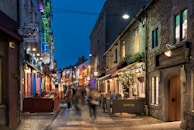 A narrow cobblestone street lined with lively cafes and shops, bathed in the soft glow of streetlights. The buildings have a charming rustic appearance with flowers adorning the window sills. People are enjoying the evening in small groups, giving the street a bustling yet cozy atmosphere.