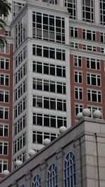 A modern building facade features a grid pattern of large windows framed with white borders, constructed with red brick and concrete materials. The structure showcases a mix of traditional and contemporary architectural elements, with ornamental spheres lining the rooftop edge.