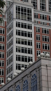 A modern building facade features a grid pattern of large windows framed with white borders, constructed with red brick and concrete materials. The structure showcases a mix of traditional and contemporary architectural elements, with ornamental spheres lining the rooftop edge.
