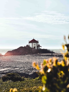 A small, white chapel with a red roof sits atop a rocky outcrop near the shoreline, with a staircase leading up to it. The beach-like foreground has a few people walking and scattered vegetation in the bottom right corner, partially out of focus. The sky is mostly clear with some scattered clouds, and the sea is visible in the background.