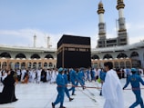 A large group of people, some dressed in blue uniforms and others in white robes, are gathered around the Kaaba during a religious pilgrimage. The Kaaba is a black cube-shaped structure located in the courtyard of a grand mosque. Two tall minarets are visible in the background along with modern construction features. The crowd appears to be moving in an organized manner, with those in blue seeming to guide or clean the area.