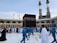 A large group of people, some dressed in blue uniforms and others in white robes, are gathered around the Kaaba during a religious pilgrimage. The Kaaba is a black cube-shaped structure located in the courtyard of a grand mosque. Two tall minarets are visible in the background along with modern construction features. The crowd appears to be moving in an organized manner, with those in blue seeming to guide or clean the area.