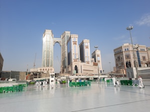Tall, modern skyscrapers with reflective glass panels stand prominently against a clear blue sky. People are walking on a large open courtyard area with white tiles. Green barriers are placed around the area, and some street lamps and cranes are visible.