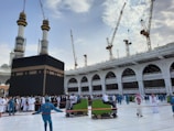 A large group of people surrounds the Kaaba in the Grand Mosque, with pilgrims wearing traditional white garments and workers in blue uniforms. There are multiple cranes visible in the background, indicating ongoing construction. The sky is partly cloudy, adding contrast to the scene.