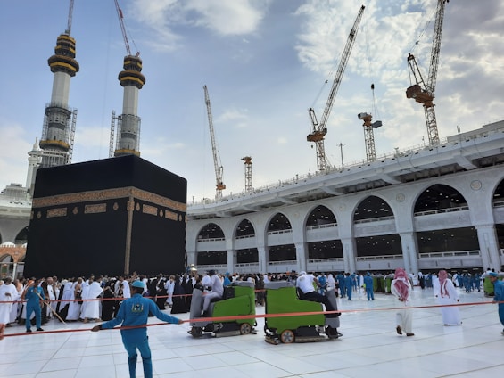 A large group of people surrounds the Kaaba in the Grand Mosque, with pilgrims wearing traditional white garments and workers in blue uniforms. There are multiple cranes visible in the background, indicating ongoing construction. The sky is partly cloudy, adding contrast to the scene.