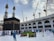 A large group of people surrounds the Kaaba in the Grand Mosque, with pilgrims wearing traditional white garments and workers in blue uniforms. There are multiple cranes visible in the background, indicating ongoing construction. The sky is partly cloudy, adding contrast to the scene.