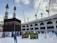 A large group of people surrounds the Kaaba in the Grand Mosque, with pilgrims wearing traditional white garments and workers in blue uniforms. There are multiple cranes visible in the background, indicating ongoing construction. The sky is partly cloudy, adding contrast to the scene.