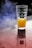 A glass with a textured surface contains a bright yellow-orange liquid resembling juice, set on a reflective surface. Smoke or mist is visible in the background, adding a dramatic effect. The glass has a label with the text 'Three Sixty Vodka.'