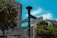 A street signpost with multiple directional signs in different languages, including English and Chinese, stands in an urban setting. The background features a tall building, lush green trees, and a bright blue sky with some clouds.