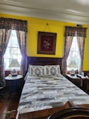 A well-decorated bedroom with vibrant yellow walls. The bed is centrally positioned with a patterned quilt and pillows. Two bedside tables with lamps flank the bed. Ornate floral curtains hang on windows on either side. A colorful painting hangs above the bed.
