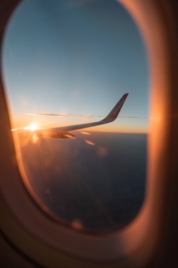 A view through an airplane window showcasing an aircraft wing with the text 'easyJet' visible, illuminated by a warm sunrise or sunset. The sky is clear with a gradient from light to dark blue, and the horizon is visible in the distance.