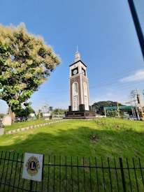 A tall clock tower stands prominently in a landscaped grassy area, surrounded by a black metal fence with a Lions International logo. The tower features a clock face on each side and a pointed top spire. A large tree with dense foliage can be seen nearby.
