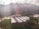 A large agricultural facility is nestled within a lush, green valley surrounded by densely forested hills. The buildings have long, rectangular roofs, likely indicating greenhouses or storage areas. Sunlight filters through scattered clouds, casting gentle rays on the landscape.