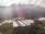 A large agricultural facility is nestled within a lush, green valley surrounded by densely forested hills. The buildings have long, rectangular roofs, likely indicating greenhouses or storage areas. Sunlight filters through scattered clouds, casting gentle rays on the landscape.