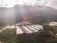 A large agricultural facility is nestled within a lush, green valley surrounded by densely forested hills. The buildings have long, rectangular roofs, likely indicating greenhouses or storage areas. Sunlight filters through scattered clouds, casting gentle rays on the landscape.