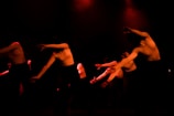 Several dancers in motion, captured under low red lighting. They are dressed in light tops and dark bottoms, performing expressive movements with their arms extended. The environment is dark, creating dramatic shadows and highlighting their dynamic poses.