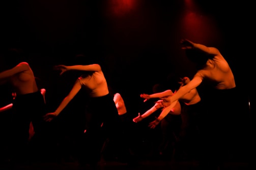 Several dancers in motion, captured under low red lighting. They are dressed in light tops and dark bottoms, performing expressive movements with their arms extended. The environment is dark, creating dramatic shadows and highlighting their dynamic poses.