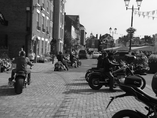 Motorcycles are parked and being ridden on a cobblestone street in a quaint town setting. The scene includes stone buildings, storefronts, and decorative street elements like hanging lights and potted plants. Several people are visible, including riders wearing helmets.