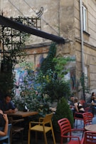 An outdoor cafe scene features several people seated at different tables amidst lush greenery and ivy-covered walls. The cafe is set against a backdrop of a worn, graffiti-adorned building with a rustic, urban atmosphere. A variety of chairs in vibrant colors are placed around wooden tables, creating a casual and eclectic vibe.