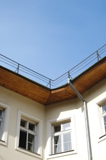A corner section of a building with a beige facade and several windows, featuring a wooden eave and a metal downspout. The sky is clear and blue above the rooftop railing.