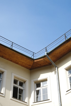 A corner section of a building with a beige facade and several windows, featuring a wooden eave and a metal downspout. The sky is clear and blue above the rooftop railing.