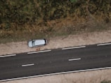 An aerial view of a road with a parked car on a dirt shoulder. A person stands beside the vehicle looking upwards, while vegetation lines the opposite side of the street.
