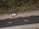 An aerial view of a road with a parked car on a dirt shoulder. A person stands beside the vehicle looking upwards, while vegetation lines the opposite side of the street.