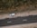 An aerial view of a road with a parked car on a dirt shoulder. A person stands beside the vehicle looking upwards, while vegetation lines the opposite side of the street.
