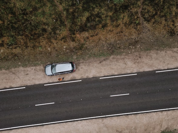 An aerial view of a road with a parked car on a dirt shoulder. A person stands beside the vehicle looking upwards, while vegetation lines the opposite side of the street.