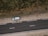 An aerial view of a road with a parked car on a dirt shoulder. A person stands beside the vehicle looking upwards, while vegetation lines the opposite side of the street.