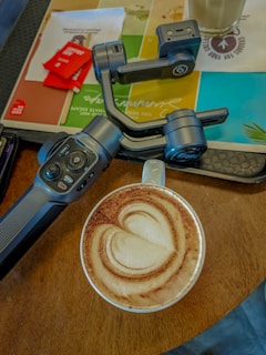 A cup of coffee with intricate latte art in the shape of a heart, placed on a wooden table. Beside the coffee, there is a handheld gimbal with a control panel featuring multiple buttons. Various napkins and sachets are visible in the background, along with a glass on a colorful placemat.