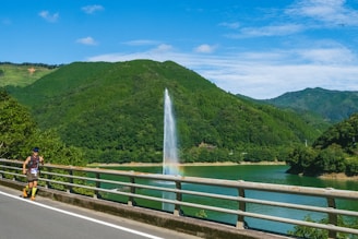 A scenic landscape featuring a lush, green mountain range under a clear blue sky with scattered clouds. A large fountain sprays water into the air, creating a visible rainbow near its base, in a lake that runs alongside a roadside. A person running along the road is visible in the foreground, wearing athletic gear and a race number.