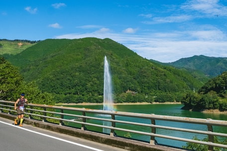 A scenic landscape featuring a lush, green mountain range under a clear blue sky with scattered clouds. A large fountain sprays water into the air, creating a visible rainbow near its base, in a lake that runs alongside a roadside. A person running along the road is visible in the foreground, wearing athletic gear and a race number.