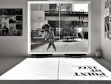 A monochrome image captures an urban scene through a large window. A person is walking outside, dressed in a checkered outfit. Inside, various posters featuring jazz musicians adorn the walls. Reflected on the interior surface is a large shadow of text advertising a jazz event.