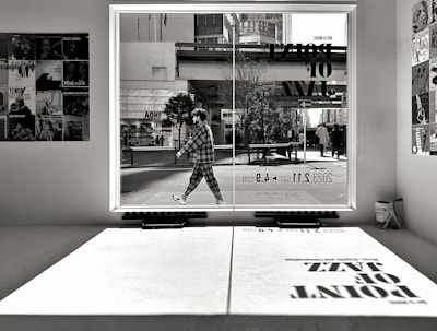 A monochrome image captures an urban scene through a large window. A person is walking outside, dressed in a checkered outfit. Inside, various posters featuring jazz musicians adorn the walls. Reflected on the interior surface is a large shadow of text advertising a jazz event.