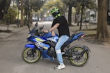 A person wearing a black shirt, light blue jeans, and a helmet is sitting on a blue motorcycle parked on a paved road. The motorcycle is decorated with colorful decals. Surrounding the scene are tall trees and a blurred urban background.