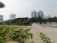 A park with a paved walkway and well-maintained green spaces is bordered by colorful blooming flowers in the foreground. Tall office buildings with distinctive architectural designs, including two with pointed rooftops, dominate the background, while trees add greenery throughout the area. People can be seen walking leisurely in the distance.