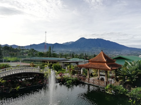 A scenic landscape with a lush garden foreground featuring a decorative pond with a central fountain. A traditional pavilion with a brown-tiled roof sits to the right, and a white arched bridge spans part of the pond. In the background, there are rolling green fields and a large mountain range under a partly cloudy sky.