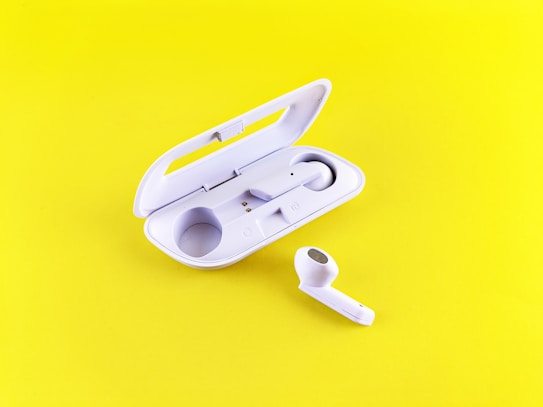 A pair of white wireless earbuds are placed on a vibrant yellow background. One earbud is seated inside an open charging case, while the other rests outside, displaying its speaker mesh.