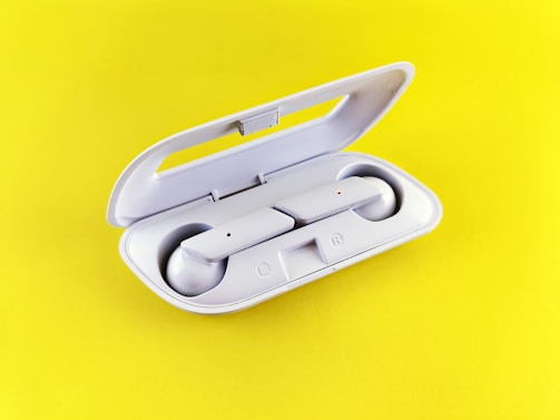 A pair of white wireless earbuds is placed inside an open charging case. The case is also white, with a smooth, sleek design, and both earbuds are fitted neatly within their respective slots. The background is a vibrant yellow, creating a strong contrast with the earbuds.