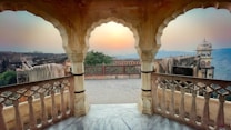 An arched, ornately designed stone balcony overlooks a serene landscape, featuring an ancient fortress with intricate carvings. The sky is a soft gradient of warm colors, indicating either sunrise or sunset, adding to the peaceful ambiance.