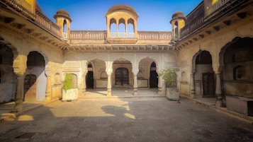 Intricately designed historical courtyard with ornate archways, columns, and open spaces. The architecture features detailed carvings and traditional elements, highlighted by warm sunlight casting shadows across the stone floor. Potted plants in corners add a touch of greenery amidst the neutral tones.
