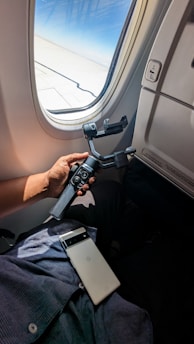 A person is holding a gimbal stabilizer in their hand while seated next to an airplane window. A smartphone rests on their lap, and the plane's wing can be seen through the window.