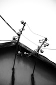 A rooftop with several electrical insulators and wires is silhouetted against a bright white sky. The stark contrast emphasizes the angular lines of the wires and insulators, giving it a geometric appearance. The texture of the roof and the materials used are evident in the image's details.
