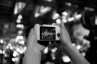 Hands holding a digital camera, with the camera's screen displaying a live scene full of lights and a crowd. The background is blurred with circular bokeh effects, creating a dynamic and lively atmosphere.