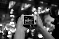 Hands holding a digital camera, with the camera's screen displaying a live scene full of lights and a crowd. The background is blurred with circular bokeh effects, creating a dynamic and lively atmosphere.