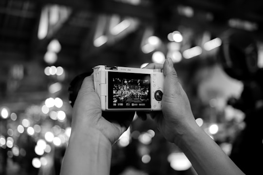 Hands holding a digital camera, with the camera's screen displaying a live scene full of lights and a crowd. The background is blurred with circular bokeh effects, creating a dynamic and lively atmosphere.